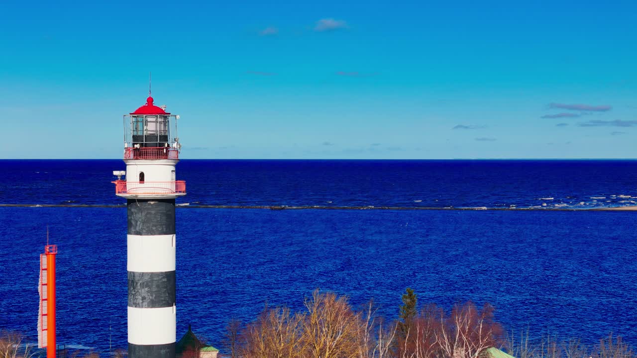 A tall black and white lighthouse rises beside bare trees and vivid blue Baltic Sea, with a clear sky and sandy coastline stretching into the horizon.