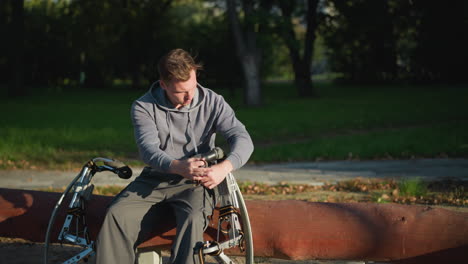 Guy with light brown hair sitting on bench outdoors wearing gray hoodie and sweatpants, focused on modified mobility device in sunny park setting surrounded by trees, sunlight