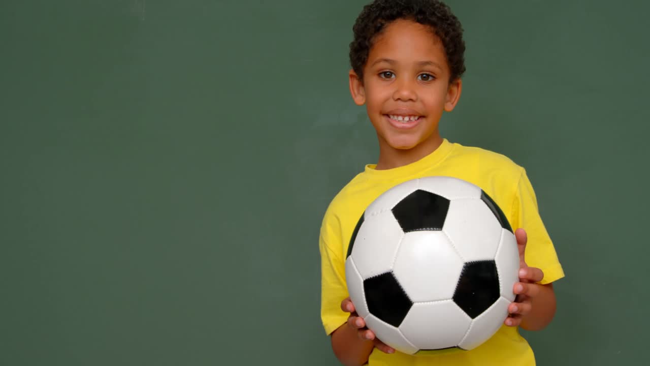 vista frontal de un escolar afroamericano de pie con el fútbol contra la pizarra en el salón de clases 4k