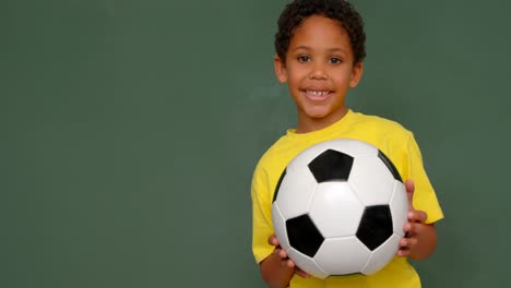 front view of african american schoolboy standing with football against chalkboard in classroom 4k