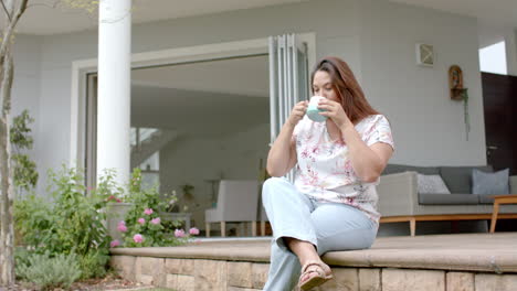 Happy-plus-size-biracial-woman-sitting-on-garden-terrace-drinking-coffee,-copy-space,-slow-motion