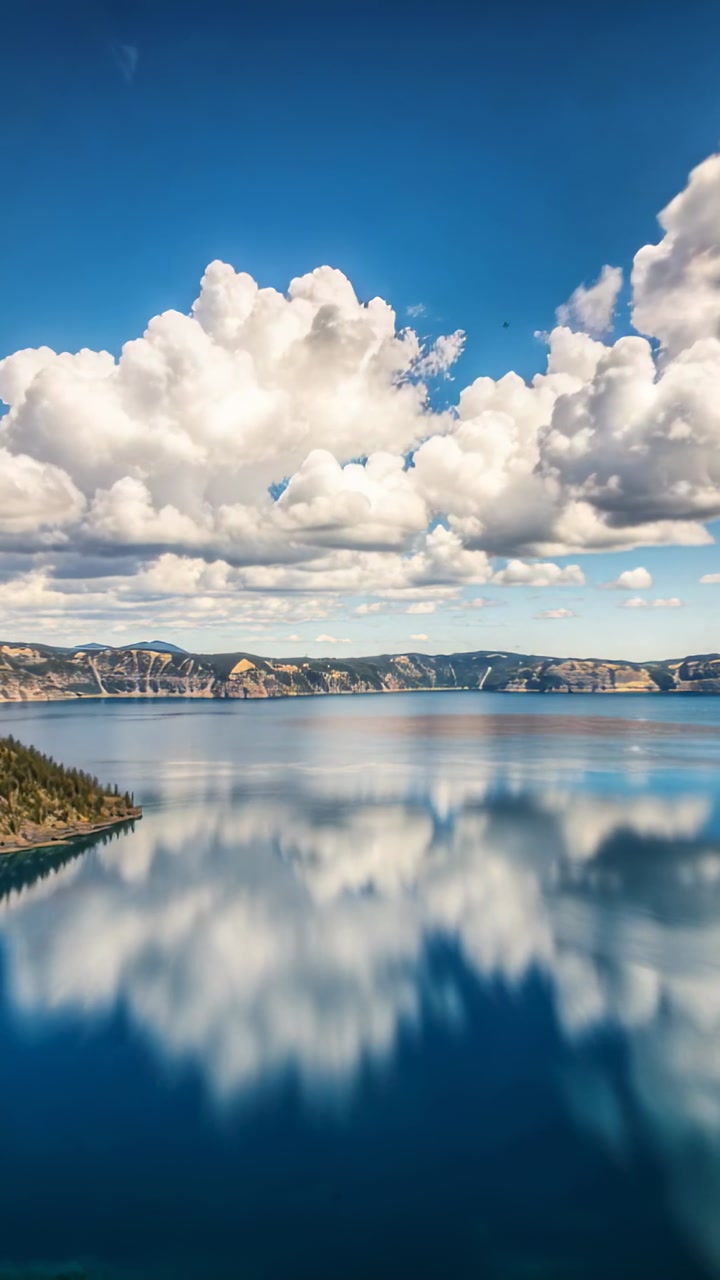 Vertical video: Drifting cumulus clouds reflecting on deep lake at shore, showing wooded peninsula