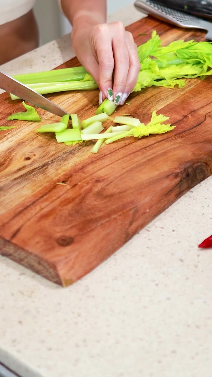 Una persona corta apio en una tabla de madera en un entorno de cocina luminosa, destacando técnicas de preparación culinaria