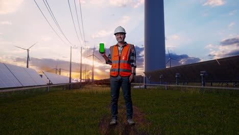 full body of asian male engineer with safety helmet smiling and showing green screen smartphone to the camera while standing with solar panel and wind turbines