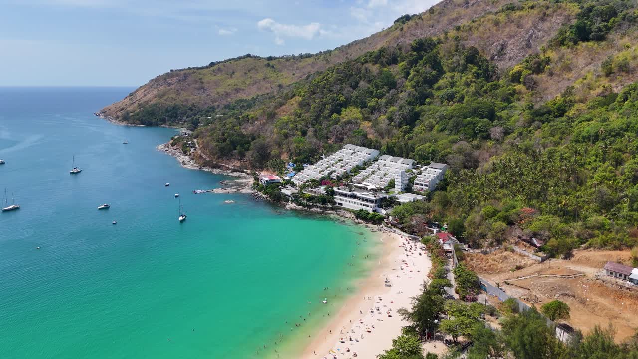 Drone footage captures Nai Harn Beach in Phuket, Thailand, with turquoise waters, lush greenery, and scattered boats under bright daylight