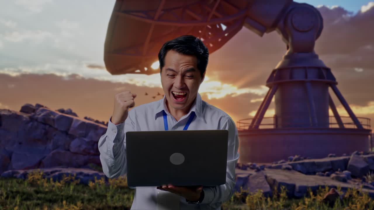 Asian Male Professional Worker Standing With His Laptop With Large Satellite Dish, He Raises His Fist Up With Screaming Goal