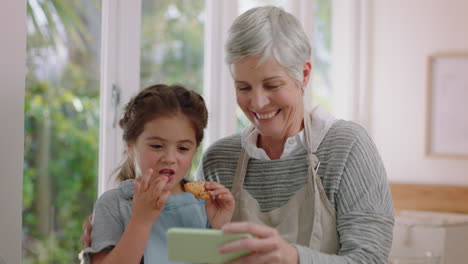 Abuela-Y-Niña-Usando-Un-Teléfono-Inteligente-Viendo-Entretenimiento-En-Línea-Comiendo-Galletas-En-La-Cocina-Nieta-Feliz-Disfrutando-Compartiendo-El-Fin-De-Semana-Con-La-Abuela-Navegando-Por-Internet-En-El-Teléfono-Móvil