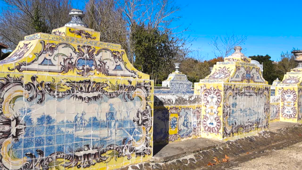 Vista próxima de um muro de jardim antigo decorado com azulejos de cerâmica tradicionais, exibindo arte ornamentada, texturas envelhecidas e luz solar quente contra um céu azul claro
