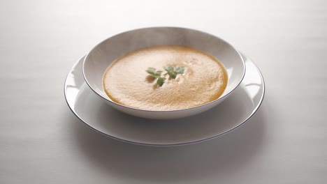 steaming hot butternut squash soup with a sprig of parsley floating on the surface, in a white bowl on a white table.