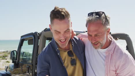 Happy-caucasian-gay-male-couple-taking-selfies-and-smiling-by-car-on-sunny-day-at-the-beach