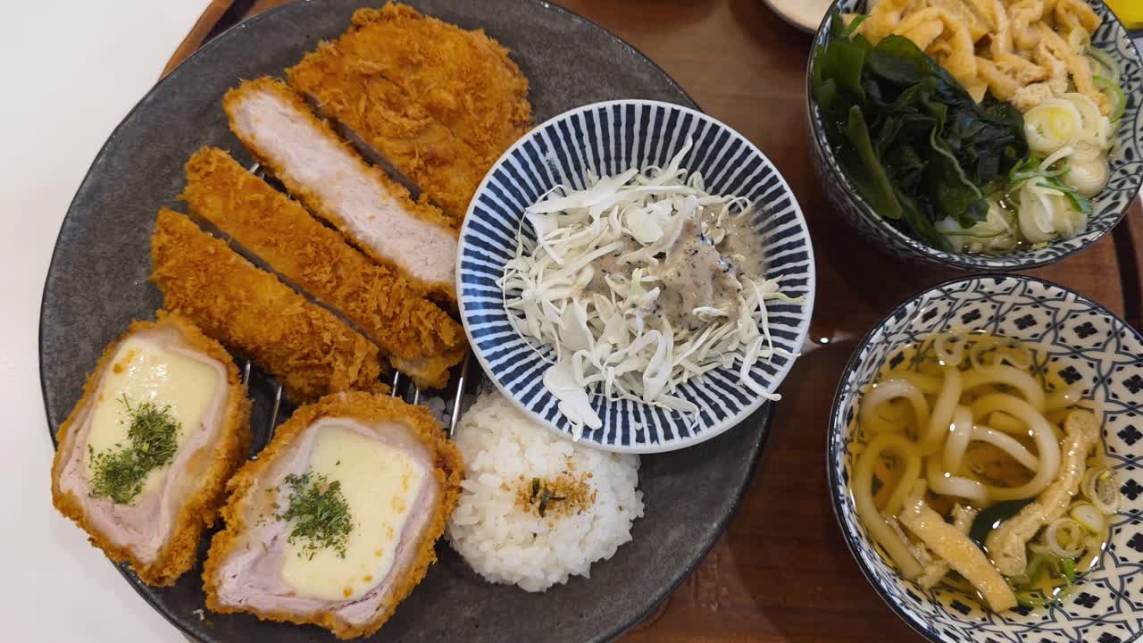 A top-down reveal of complete Japanese tonkatsu set meal, featuring crispy original and cheese-filled pork cutlets, rice, shredded cabbage salad, and a bowl of udon noodle soup