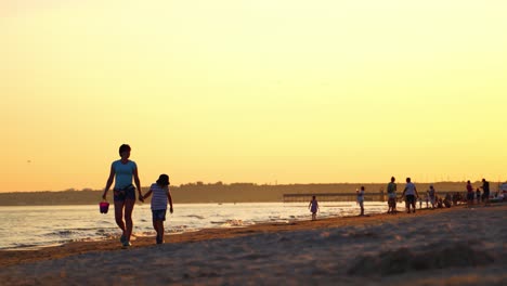 Mother with son are walking near the sea at sunset. Sealine with people in the evening in summer. Enjoyable vacation at the seaside.