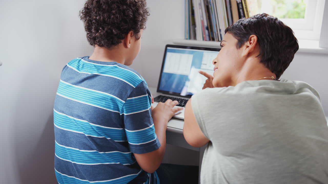 vista trasera de la madre ayudando al hijo usando la computadora portátil en el escritorio en el dormitorio
