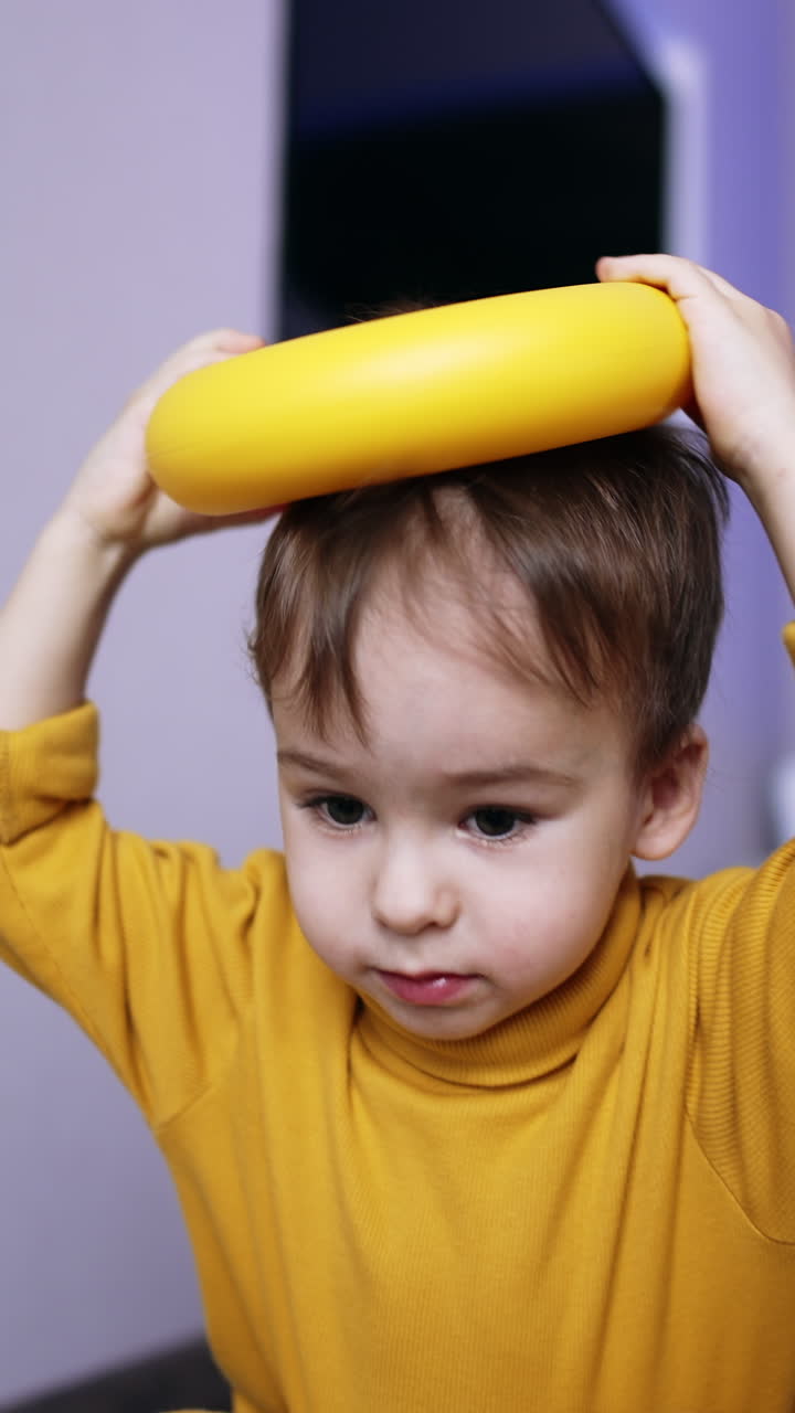 Beautiful brunet male kid in yellow sweater plays with toys. Toddler places ring from toy pyramid on his head and drops it. Close up. Vertical video
