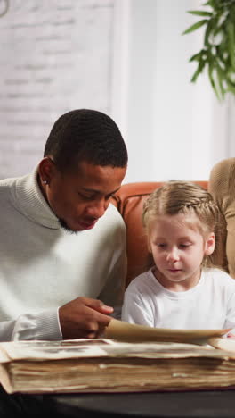 african-american man shows photo of grandparents to little girl in album sitting with blonde wife on sofa in cozy living room slow motion