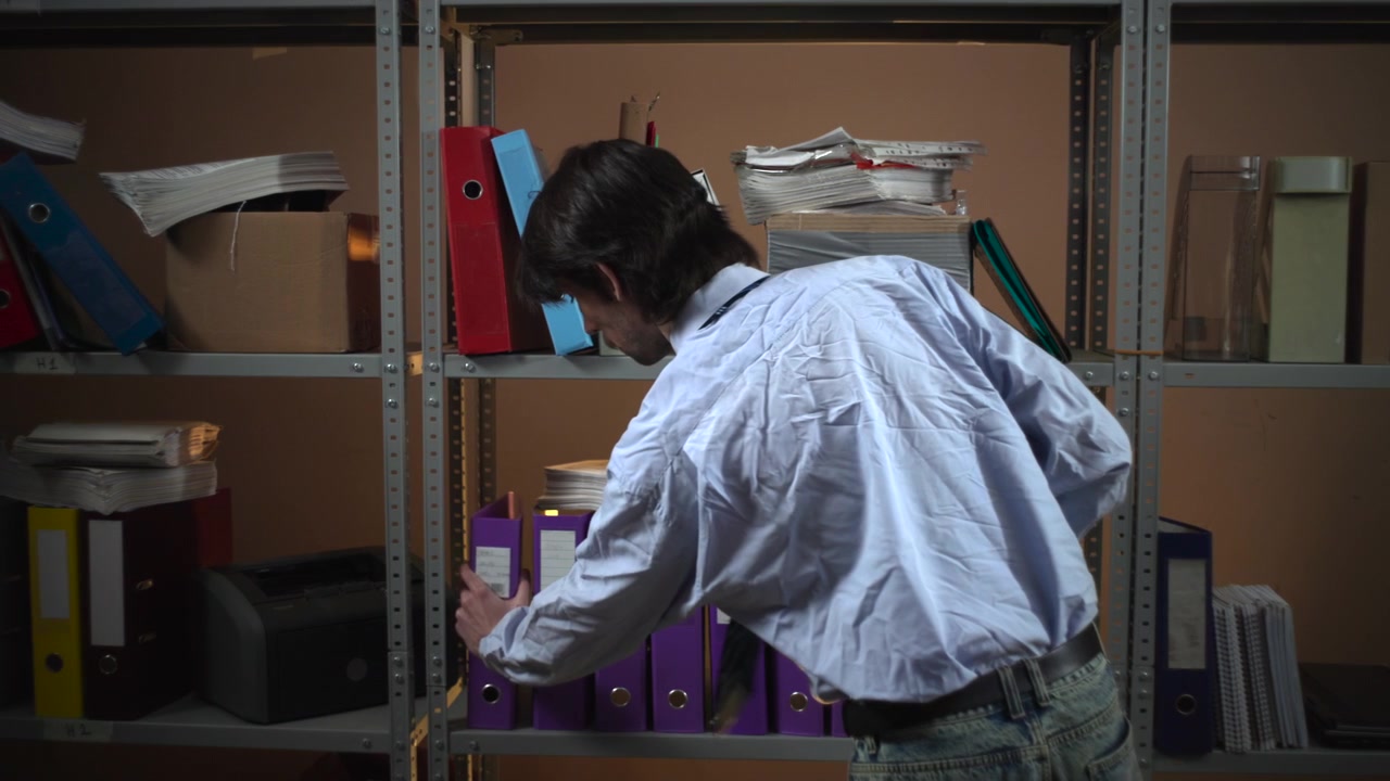 Interior de escritório com pouca luz, com prateleiras repletas de pastas, arquivos e documentos, enquanto um homem está por perto segurando uma caneca e uma pasta.