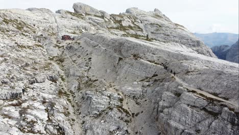 An-aerial-drone-shot-of-a-group-of-hikers-walking-towards-a-house-on-a-mountain-trail-on-a-steep-cliff-with-the-amazing-landscape-of-the-Dolomites-in-the-background-in-North-Italy