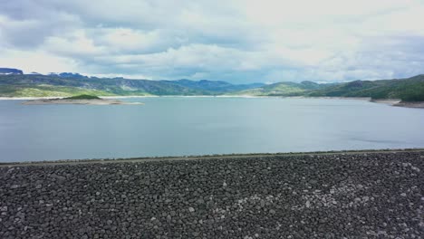 Massive-water-magazine-Sysen-dam-in-Hardangervidda-Norway---Aerial-flying-backwards-over-edge-of-dam---From-lake-overview-to-full-dam-overview