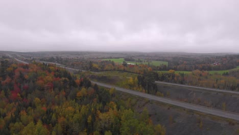 Eine-Luftaufnahme-Einer-Autobahn-Mit-Autos-Darauf-Neben-Einem-Atemberaubenden-Wald-Voller-Herbstfarben-Und-Einem-Großen-Fluss,-Der-Daneben-Fließt