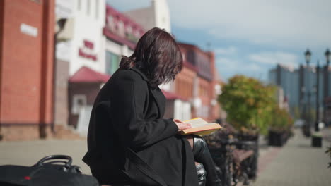 young girl in black coat seated outdoors, focused on reading her book, hair flows with the wind, urban buildings and lamp poles in blurred background