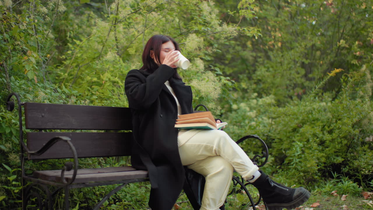 Forest visitor in long jacket, white trousers and black boots sits on bench, reading novel and flipping pages, smiling between sips of tea from paper cup, autumn arboretum around with soft greenery