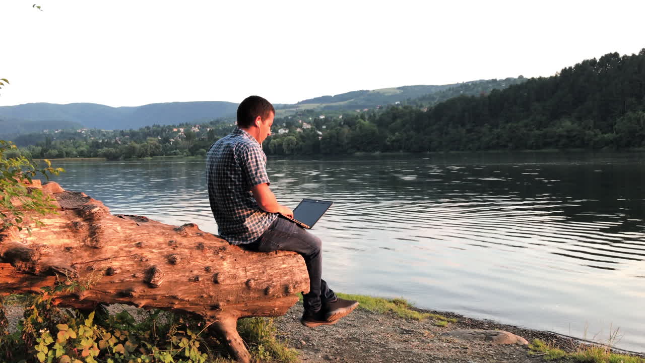 Un uomo lavora in smart working da remoto nella natura con un laptop, seduto su un ceppo d'albero e che guarda un lago.