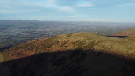 Early-sunlight-on-highland-Moel-Famau-mountain-peak-aerial-rising-view-across-vast-frosty-idyllic-farmland-countryside