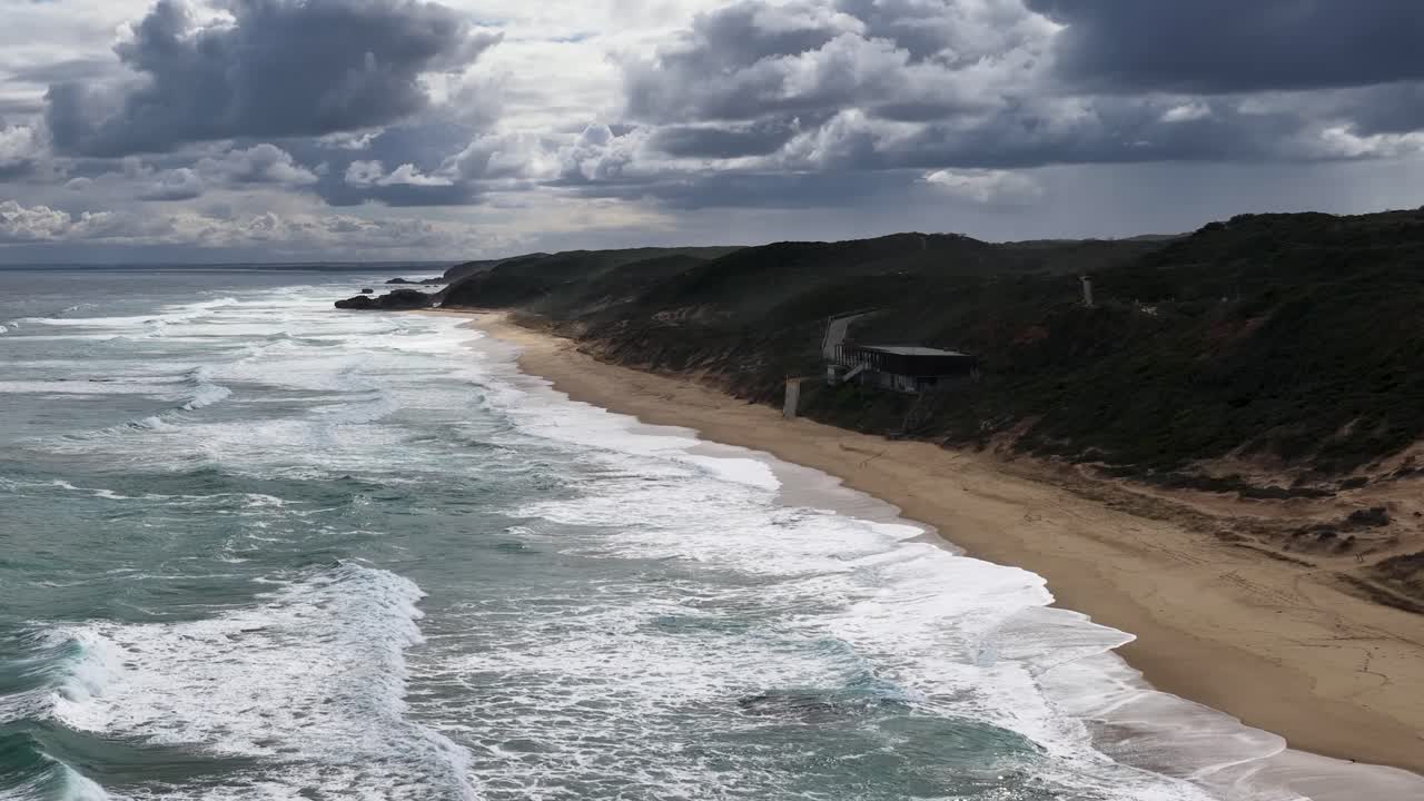 Drone camera glides above Portsea coastline, capturing dramatic waves, sandy shore, and moody sky