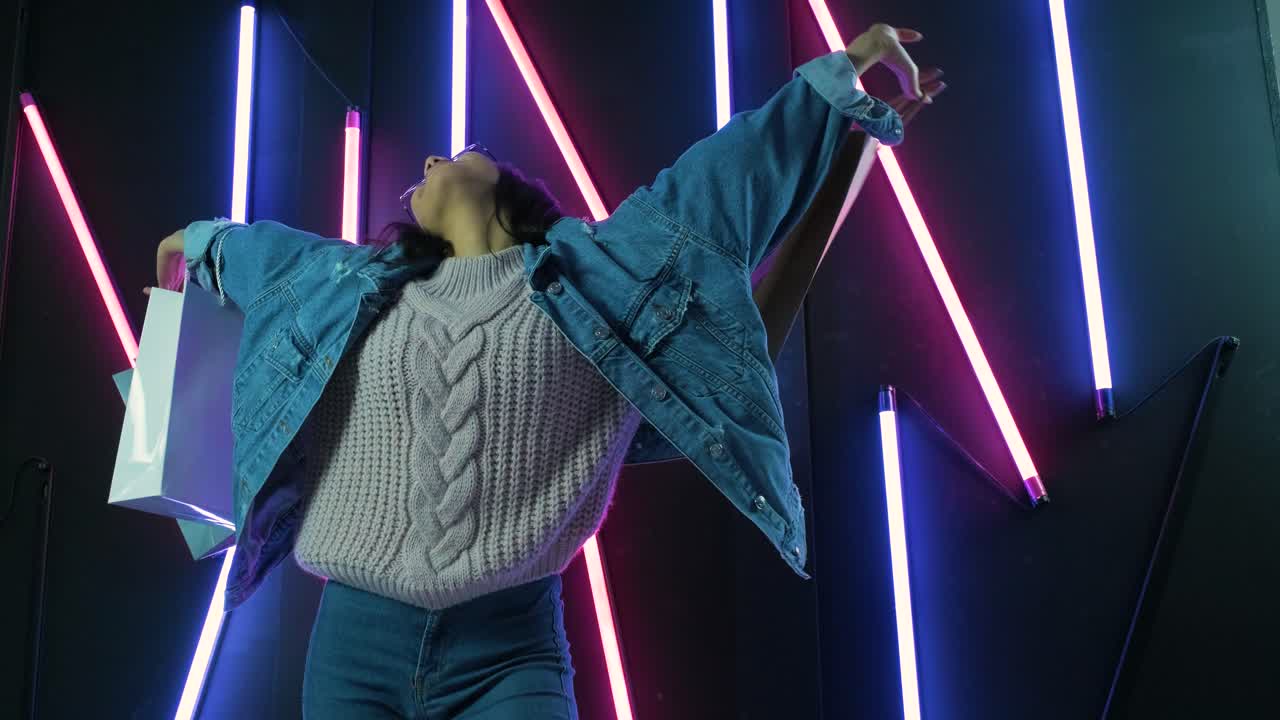 joven con chaqueta de vaqueros azul con bolsas de compras en el fondo de lámparas de neón.