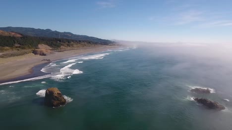 aerial: flying over the oregon coastline the waves crash against the shore as the fog rolls in