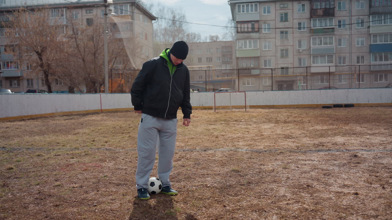 진흙 투성이 경기장에서 축구공을 저글링하는 백인 남성 프리스타일 선수, 회색 하늘, 아파트 배경, 초록색 후드티와 비니, 집중된 발놀림, 개인 훈련 세션, 리듬감 있는 공 컨트롤