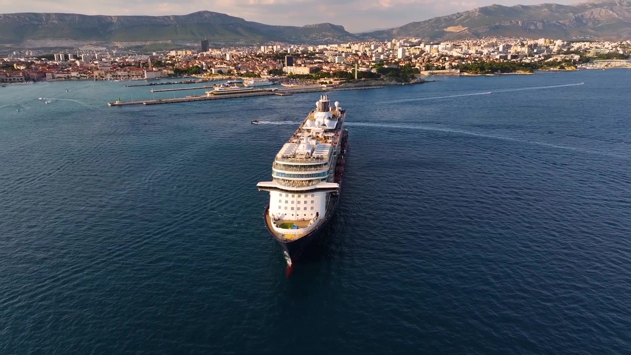 Krydstogtskib sejler ind i havn, mens kystlinje og by dukker op i baggrunden. Passagerer forbereder sig på at gå fra borde i en travl turistdestination