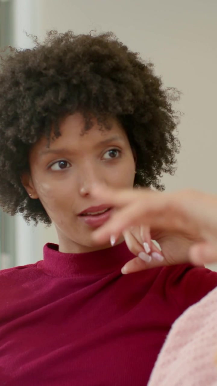 Vertical video: Holding phone, woman in burgundy gesturing on sofa, listening to friend, copy space