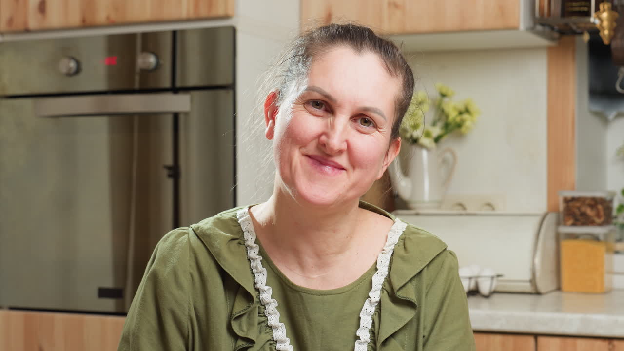 Portrait of cheerful woman with kind expression tilting head and smiling gently in cozy kitchen filled with soft light, wooden cabinets, flowers, and warm atmosphere suggesting comfort and happiness