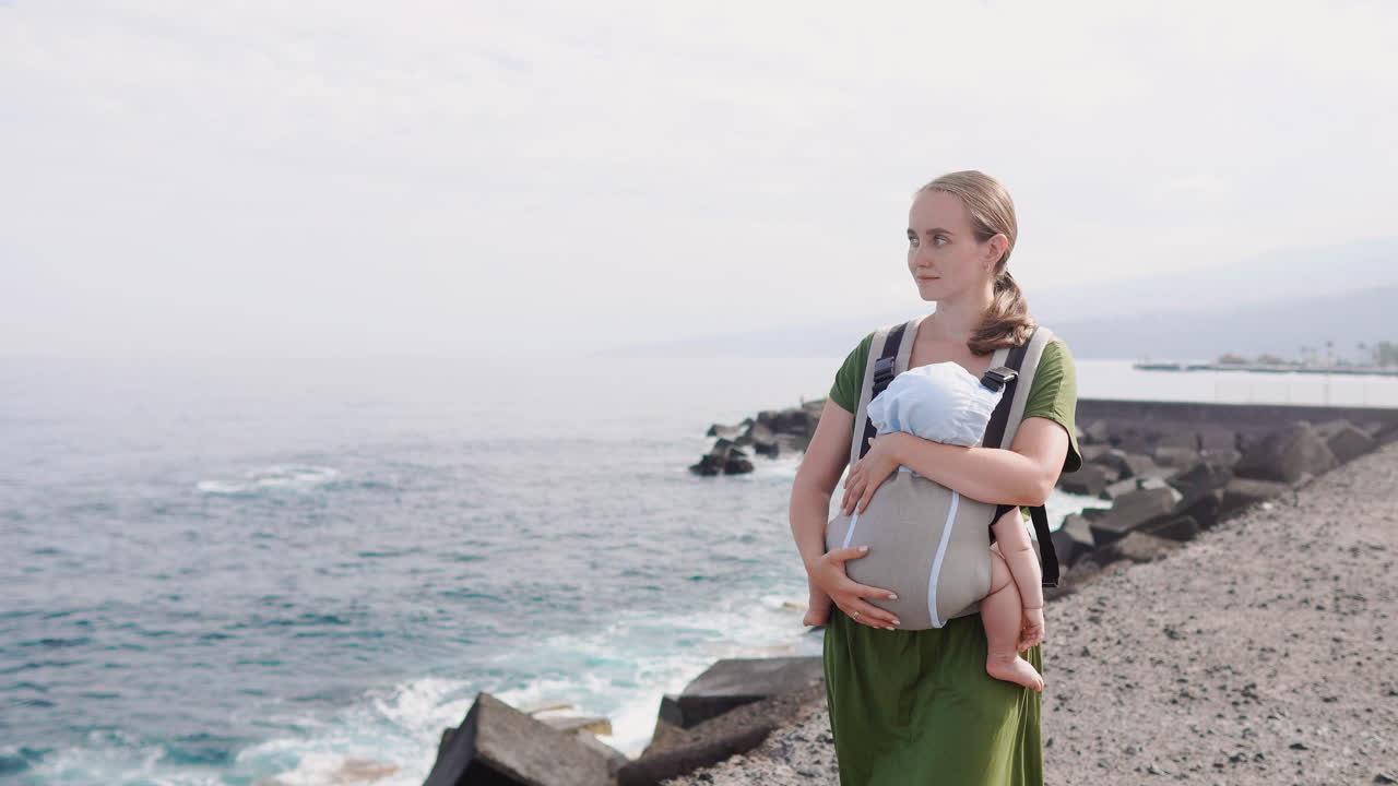 On a sunny summer day by the sea, a mother and baby enjoy a happy walk amidst nature's beauty. The child is snug in a kangaroo backpack