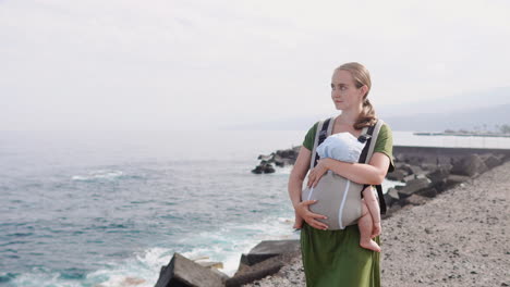 On a sunny summer day by the sea, a mother and baby enjoy a happy walk amidst nature&#039;s beauty. The child is snug in a kangaroo backpack
