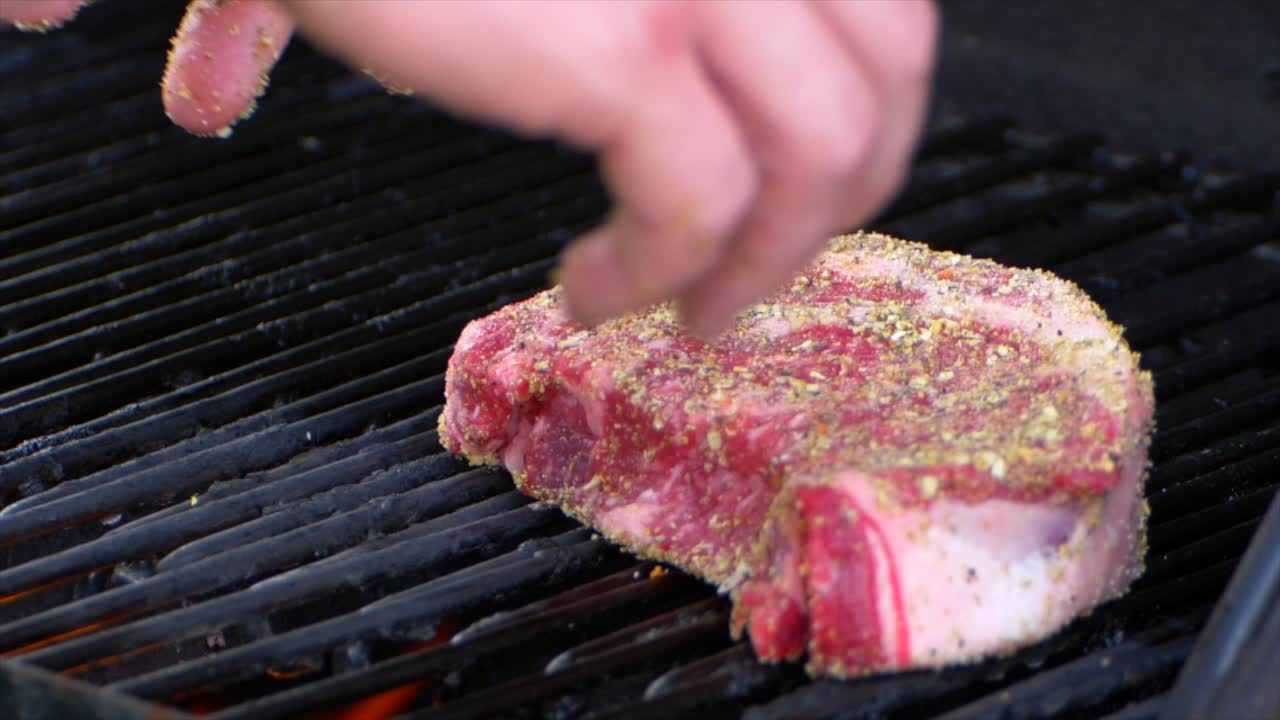 두 손으로 두꺼운 생 립아이 스테이크를 그릴에 놓고 굽는 과정을 슬로우 모션으로 시작합니다.