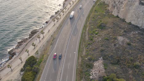 The-scenic-coastal-road-winds-its-way-along-the-breathtaking-cliffs,-with-cars-bustling-by-and-the-majestic-sea-stretching-out-on-the-side