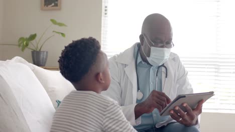 Senior-african-american-male-doctor-using-tablet-talking-to-boy-patient-at-home,-slow-motion