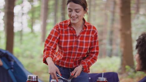 group of female friends on camping holiday in forest cooking meal sitting by tent together