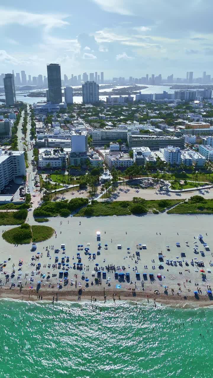 un dron vuela lateralmente sobre el océano atlántico en modo retrato, capturando las olas del océano, los bañistas, la ciudad de miami beach, y el horizonte de miami en el fondo
