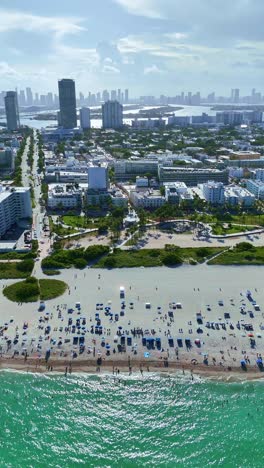 a drone flies sideways over the atlantic ocean in portrait mode, capturing ocean waves, beachgoers, miami beach city, and the miami skyline in the background