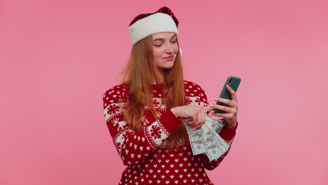 Girl in red christmas sweater using mobile phone hold money dollar cash doing online gift shopping