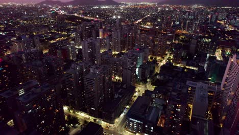 Illuminated-futuristic-night-time-Santiago,-Chile-high-rise-cityscape-aerial-view-flying-over-diverse-old-and-modern-skyscraper-capital