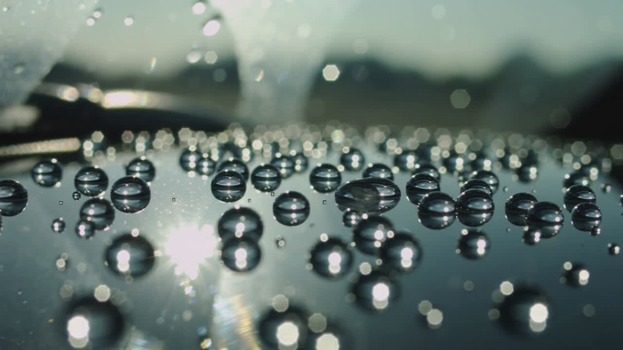 Shimmering droplets shifting and merging on car hood near windshield while sun reflection moving