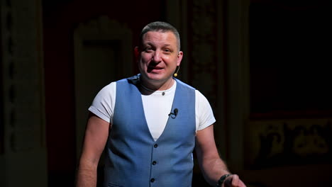 performer announces future theater season. actor is sitting in empty theater on red velvet chair with sheet of paper in hands.