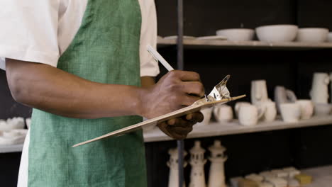 primo piano di un impiegato maschio americano irriconoscibile che scrive su una lavagna per appunti e fa un inventario delle ceramiche nel negozio di ceramiche