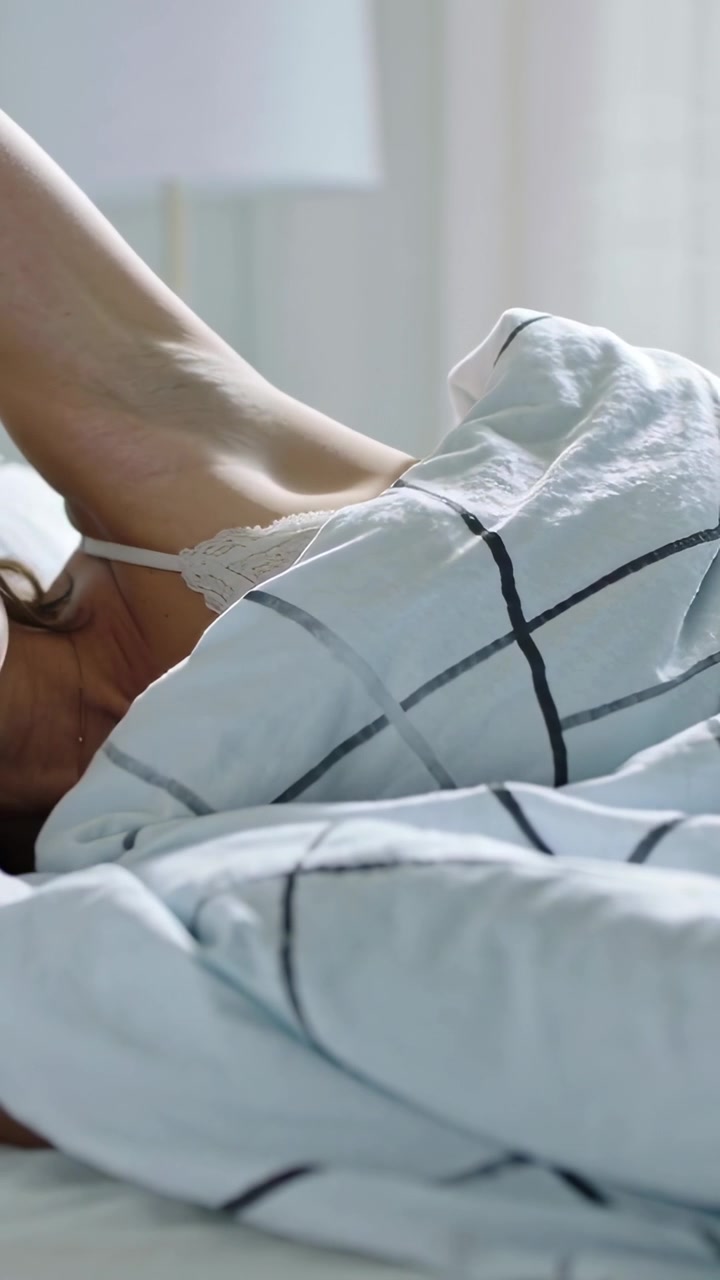 Vertical video: Waking woman pulling duvet, stretching arm in bedroom, in camisole to morning light