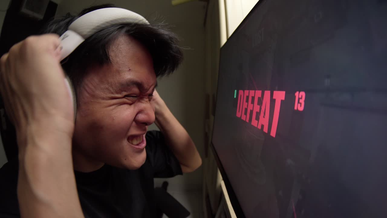 Seorang pria muda Asia mengenakan headphone bereaksi dengan frustrasi terhadap pesan kekalahan di monitor komputernya saat bermain game.