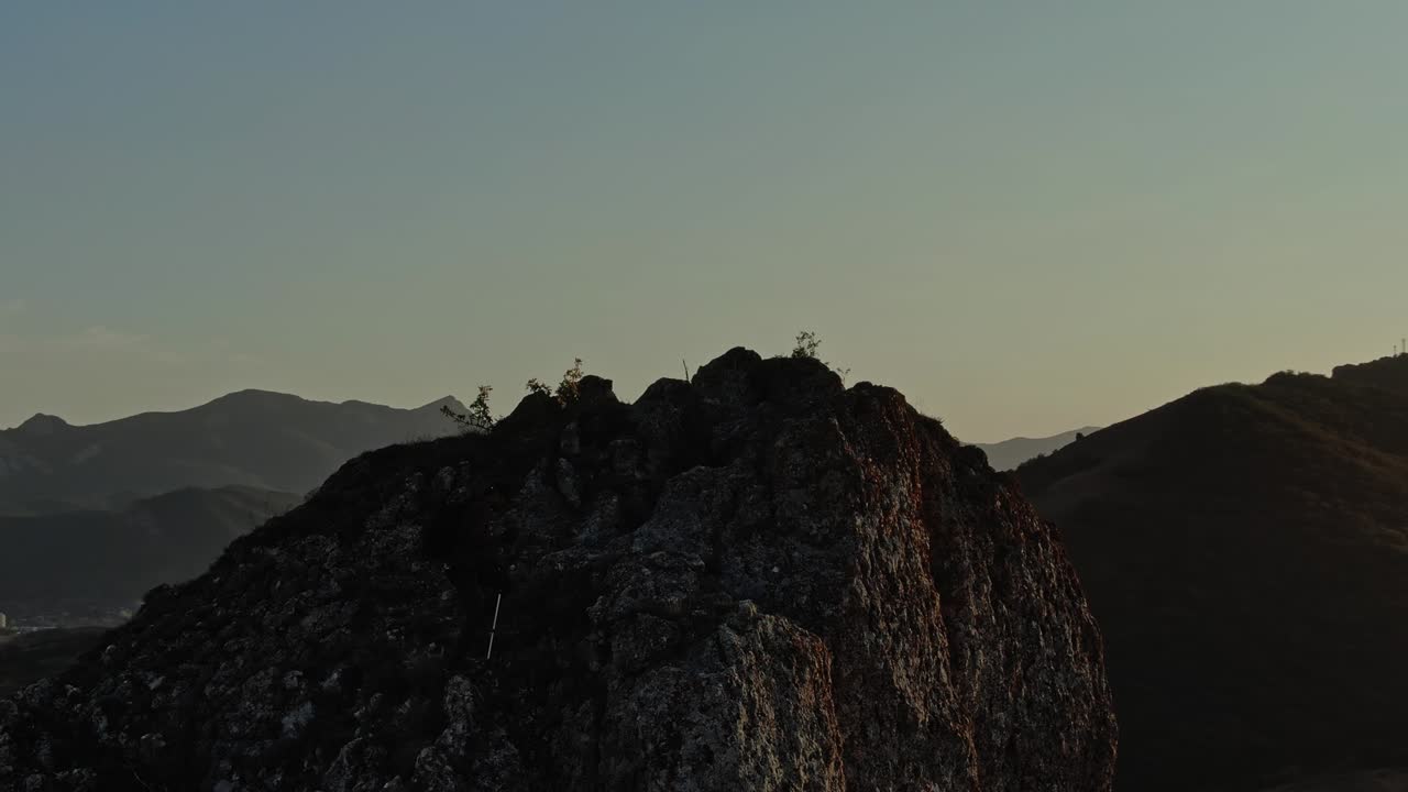 Hiker in Baseball Cap with Backpack and Nordic Walking Sticks Climbs a Rocky Top of a Mountain on the Background of a Mountain Landscape and Stands in the Rays of Sunset. Aerial view. Surveying by drone with flying in a circle.